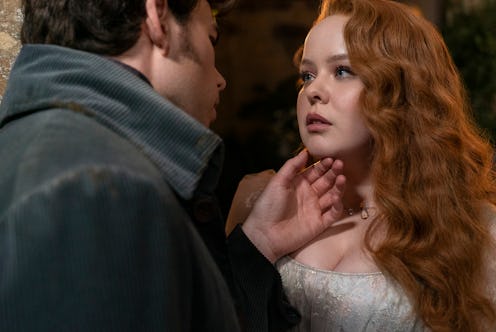 A woman with curly red hair, looking intently at a man who gently holds her chin, both illuminated s...