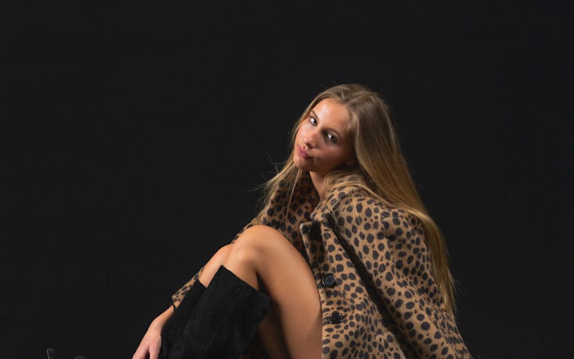 A woman sitting on the floor in a leopard print outfit and black boots, looking over her shoulder ag...