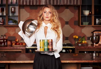 A woman in a white blouse and black skirt holds a tray with food cans and a lifted lid in a kitchen ...