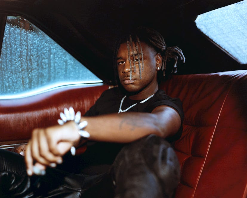 Man with braids wearing a black shirt and silver jewelry, sitting in a red car interior, looking int...