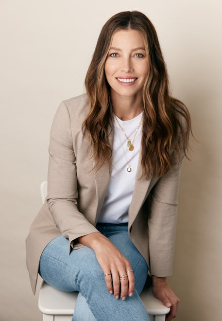 Jessica Biel wearing a tan blazer, white t-shirt, layered necklaces and jeans. She sits against a be...