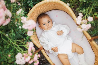 A baby in a vintage outfit lies in a bassinet outdoors.