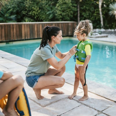 A mom snaps a life jacket on her daughter by the pool.