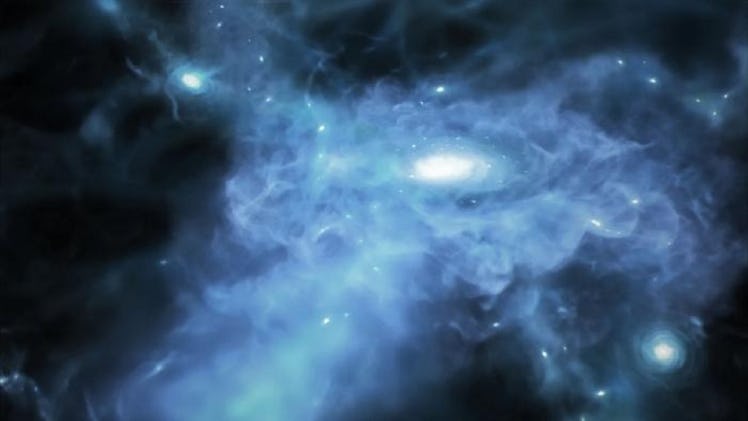 image of a small round galaxy in the middle of a cloud of blue and white gas, in space.