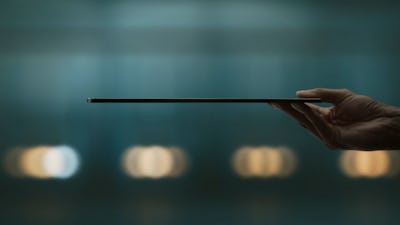A hand presents the side profile of an ultra-thin laptop against a blurred blue background with soft...