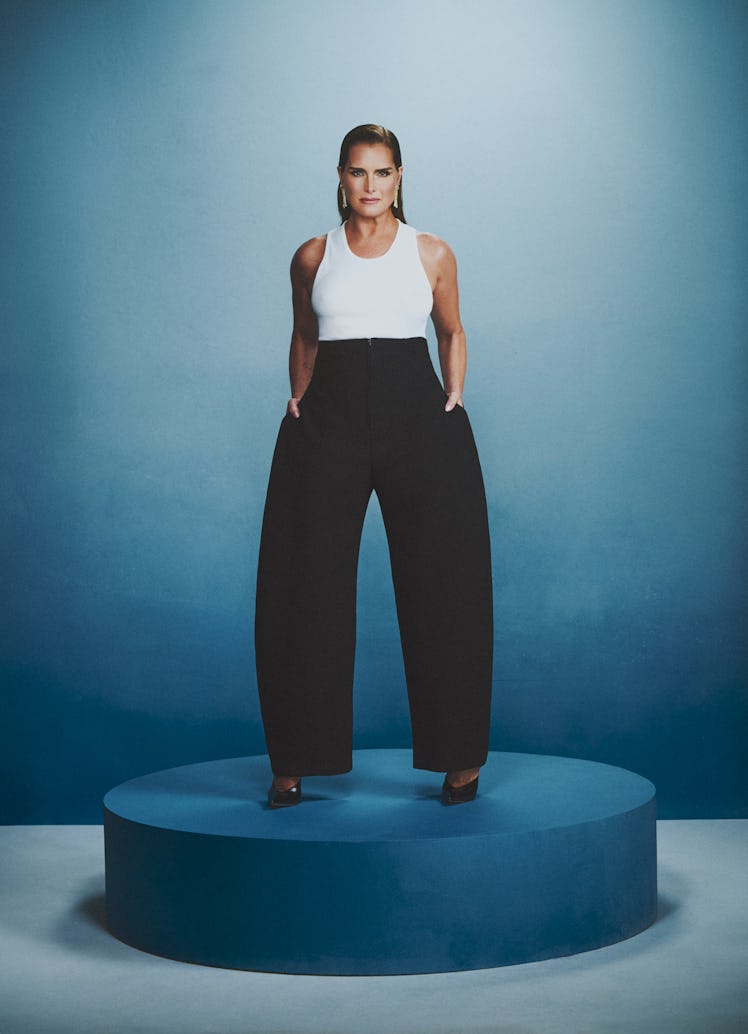 A woman stands confidently on a blue pedestal, wearing a white top, oversized black pants, and heels...