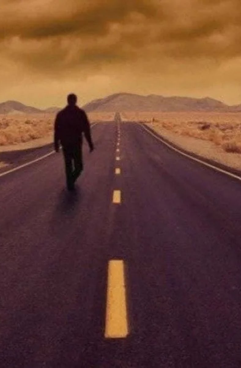 A person walking down a deserted road with yellow skies and a "slippery when wet" sign to the side.