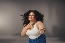 Michelle Buteau smiling and pointing wearing a white tank top and jeans against a gray background.