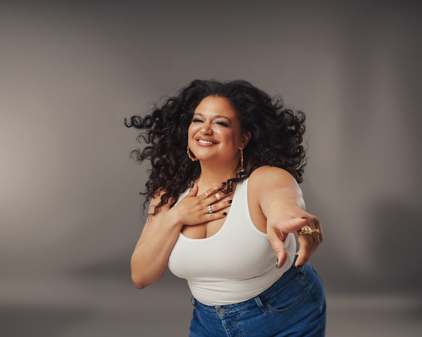 Michelle Buteau smiling and pointing wearing a white tank top and jeans against a gray background.