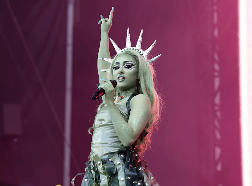 Woman performing on stage wearing a crown and an ornate costume, raising one hand, with a pink stage...