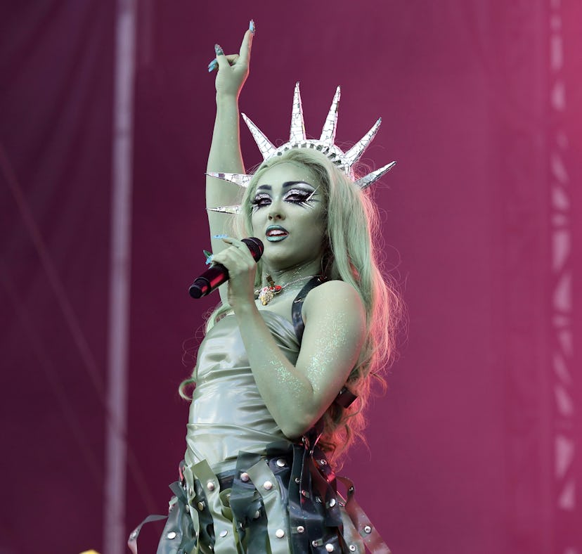 Woman performing on stage wearing a crown and an ornate costume, raising one hand, with a pink stage...