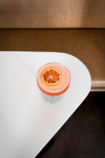 A glass of pink smoothie topped with a slice of dried orange, set on a white curved table with a cop...