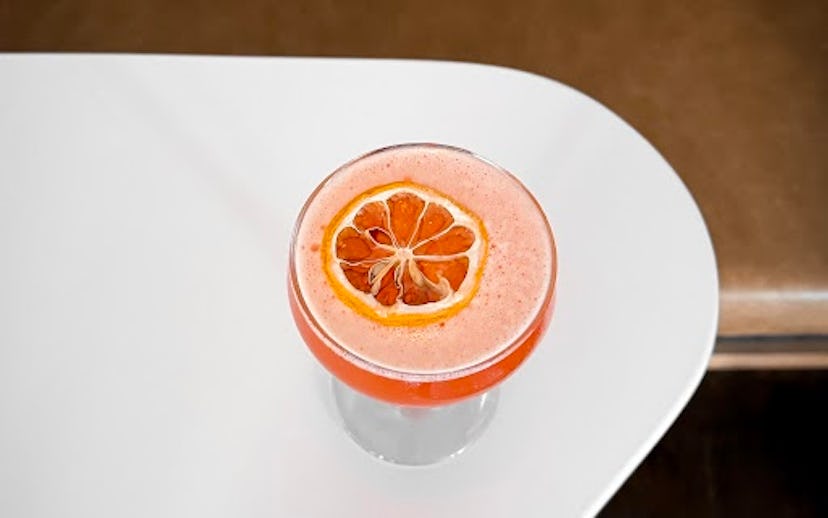 A glass of pink smoothie topped with a slice of dried orange, set on a white curved table with a cop...