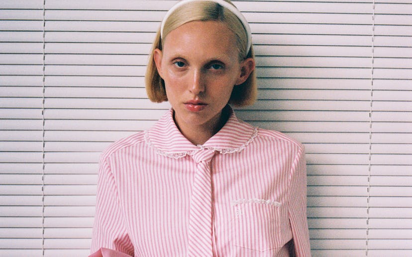 Woman in pink striped shirt and pleated skirt sitting against a blind-covered background.