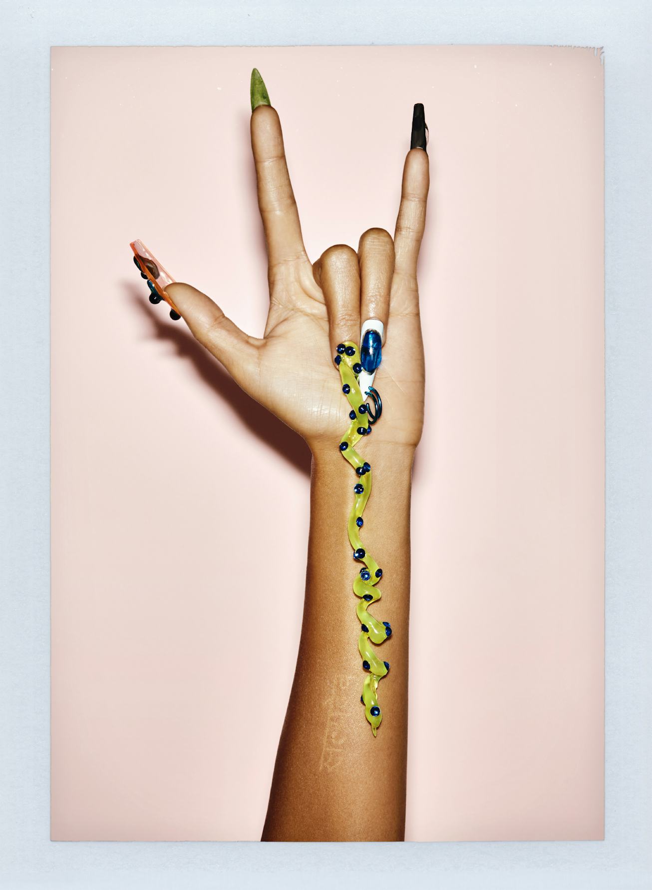 A hand against a pink background making a rock n' roll sign, adorned with multicolored, long fingern...