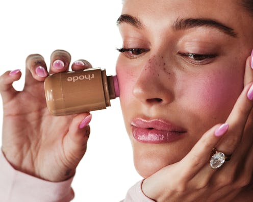 A woman with freckles examines a bottle of foundation, holding it close to her face with manicured h...
