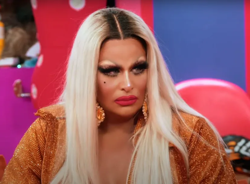 Drag queen with blonde hair and meticulous makeup wears a shimmering golden outfit, looking pensive ...