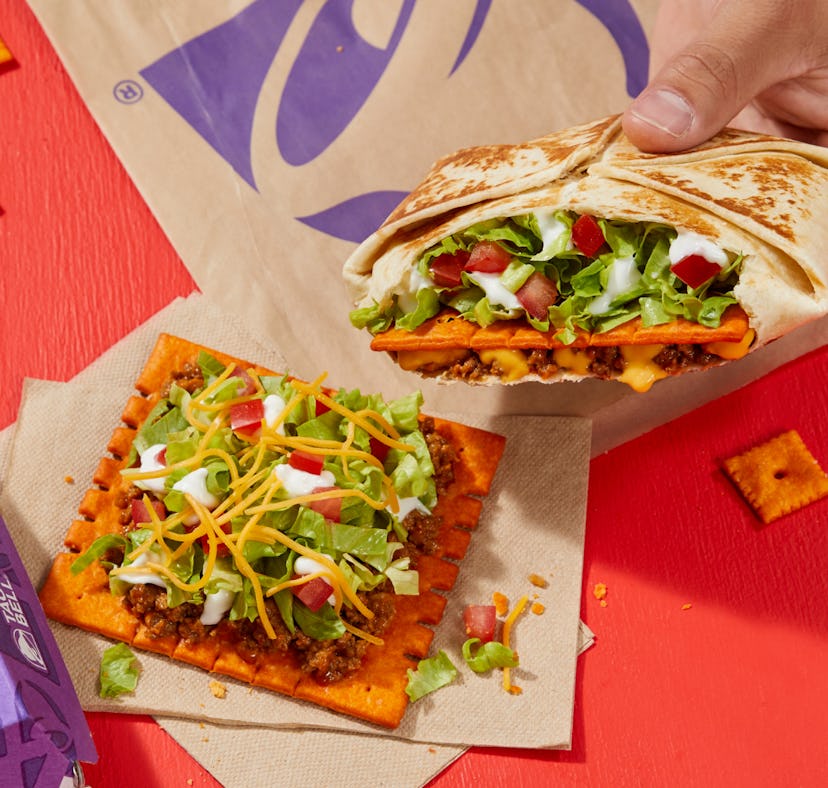 Hand holding a taco next to a square, crispy snack topped with cheese and vegetables on a red table.