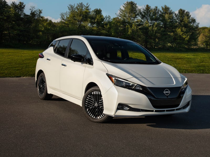 A white Nissan Leaf parked on a paved area with a backdrop of trees under a clear sky.