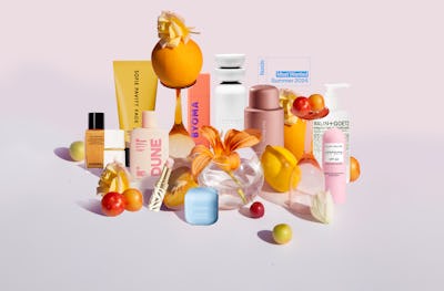 An array of colorful beauty products displayed with vibrant flowers and tropical fruits on a soft pink background.