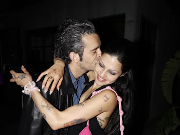 A man in a black leather jacket kissing a smiling woman with black hair on the cheek at night. Both ...