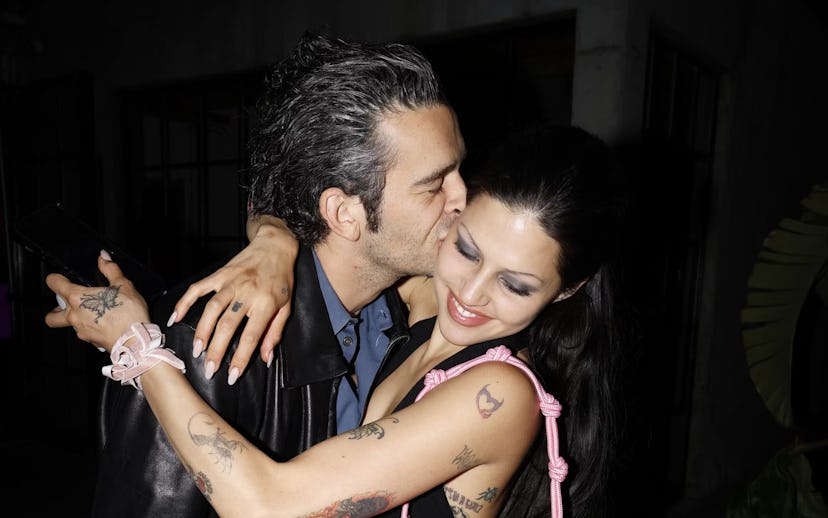 A man in a black leather jacket kissing a smiling woman with black hair on the cheek at night. Both ...