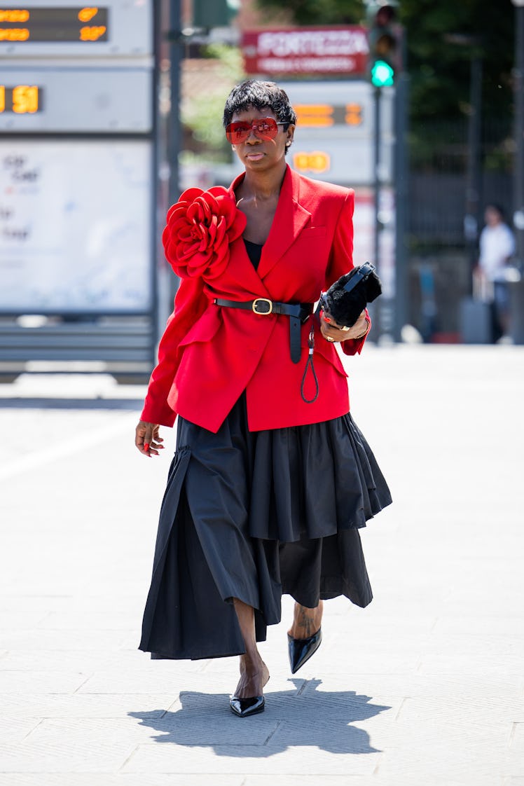 A guest wears red belted blazer, black asymmetric skirt, heels, sunglasses, bag during Pitti Immagin...