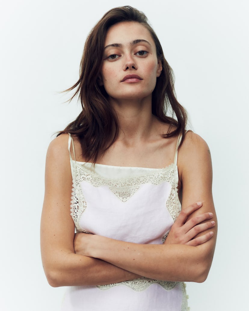 Portrait of a woman with arms crossed, wearing a white lace-trimmed top, against a plain background.