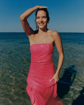 Woman in pink dress smiling by the sea, hand on head, clear blue sky and water in background.