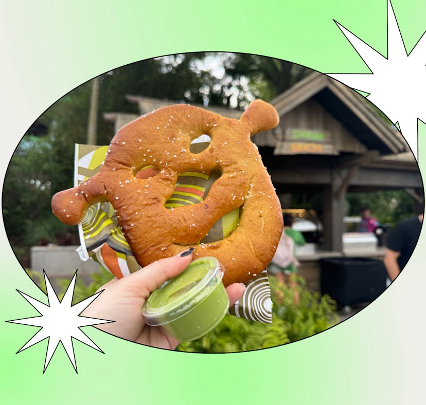 A hand holding a pretzel shaped like a Mickey Mouse head with a container of dipping sauce, in front...