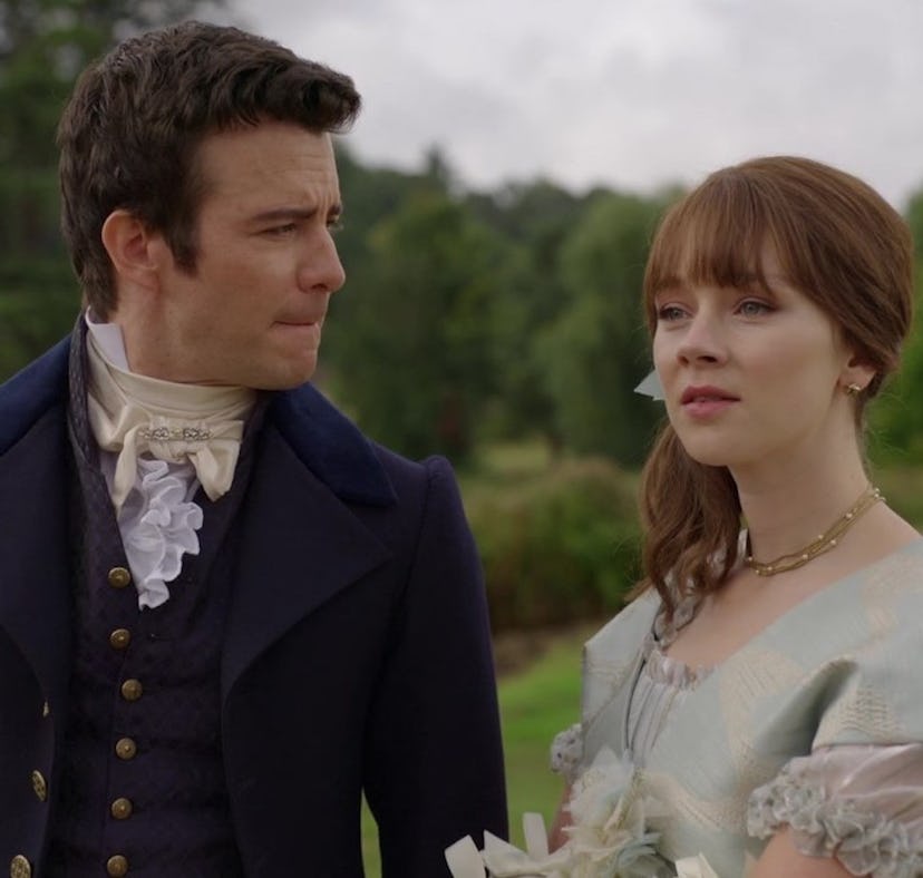 A man and a woman in historical clothing walk outdoors, seemingly in conversation, with a lush green...