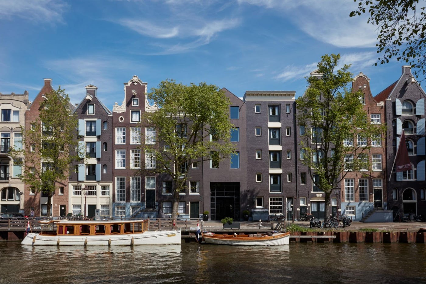 Traditional Dutch buildings line a calm canal with boats moored along the side on a sunny day in Amsterdam.