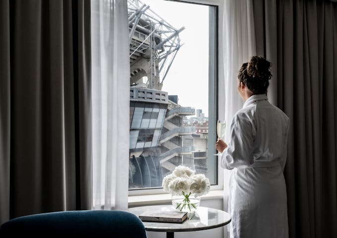 A woman in a bathrobe stands by a window, holding a drink, looking out at a large stadium structure.