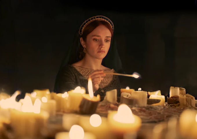 A medieval woman in a headdress lights a candle among many on a table, illuminating her focused expression.