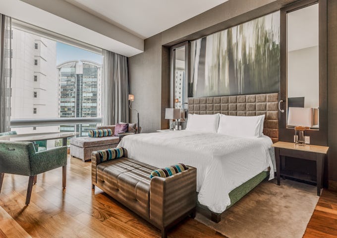Modern hotel room with a large bed, sleek furnishings, and a city view through large windows.