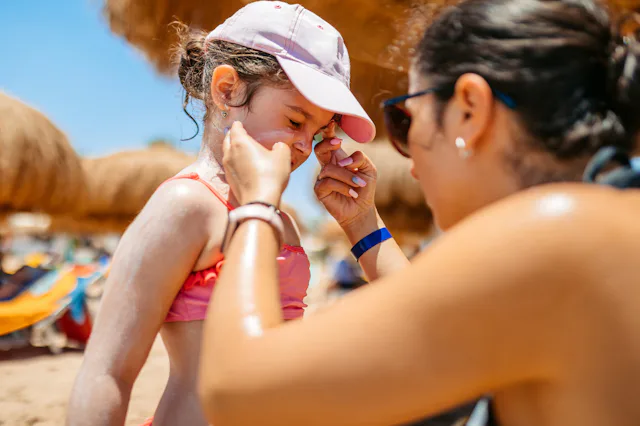 A woman applies sunscreen on a girl's nose at a sunny beach, both wearing summer clothes and hats.
