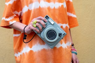 Person in an orange tie-dye shirt holding a vintage-style camera against a yellow wall.
