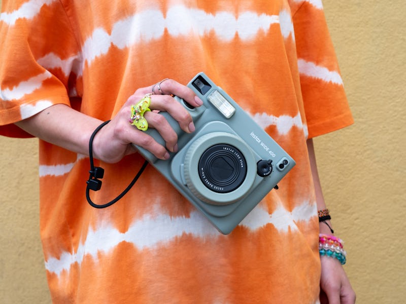 Person in an orange tie-dye shirt holding a vintage-style camera against a yellow wall.