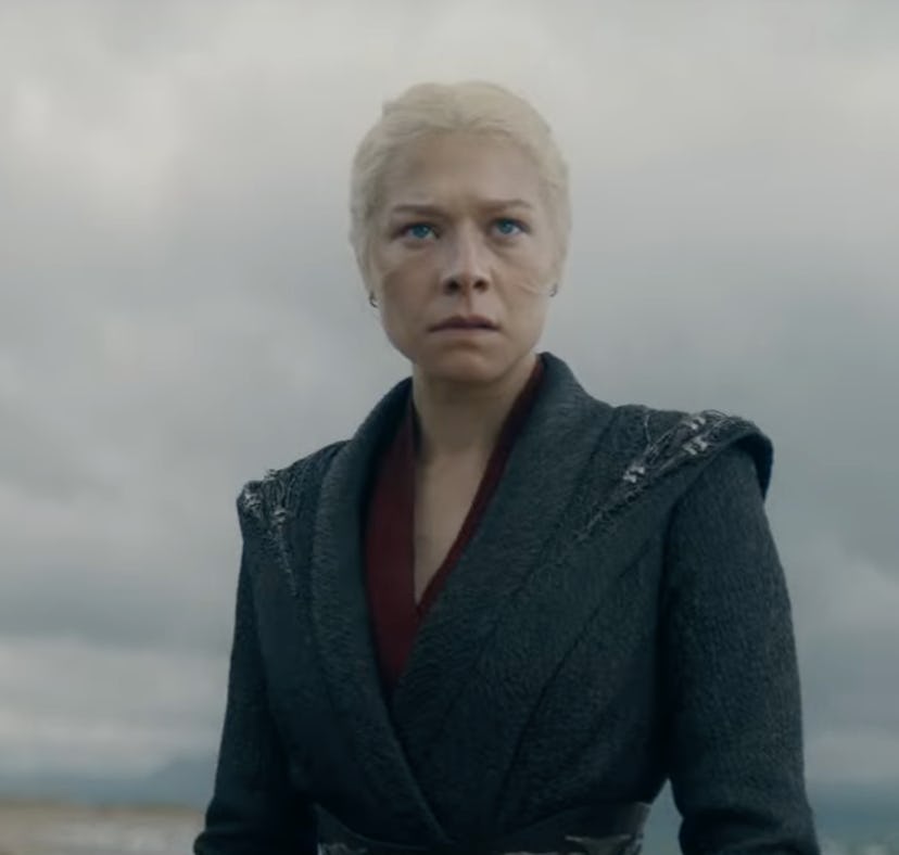 A woman with platinum blonde hair, dressed in dark clothing, stands solemnly with a cloudy sky behin...