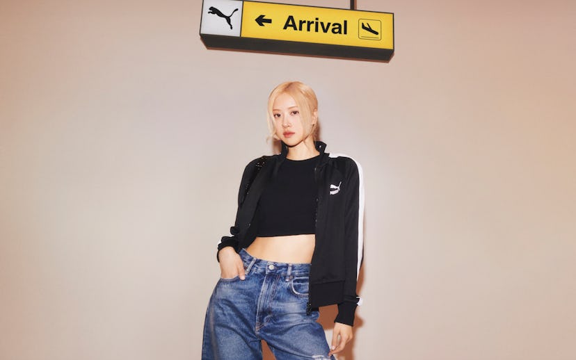 A young woman in a cropped black hoodie and jeans stands under an "Arrival" sign with a Puma logo.