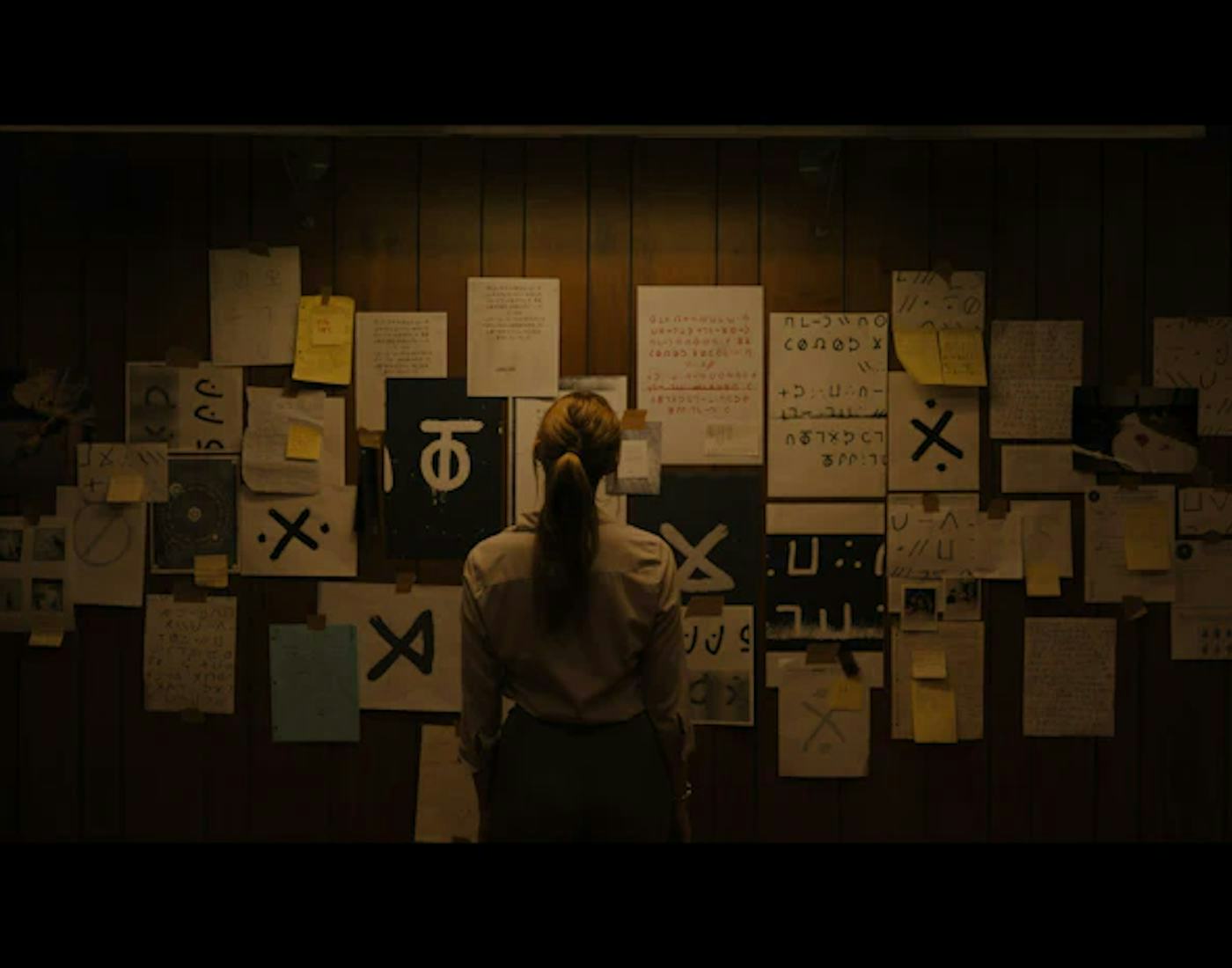 Woman standing in front of a wall covered with various mysterious symbols and notes, in a dimly lit room.