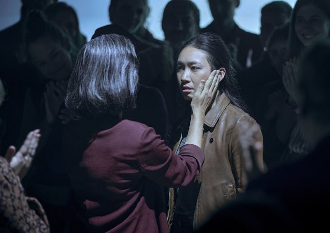 An emotional scene where a woman comforts another by touching her face, surrounded by a group of people in a dimly lit room.