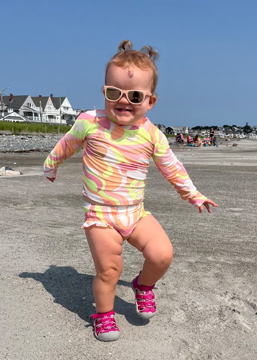 A photograph of a smiling toddler wearing sunglasses and a bathing suit at the beach.