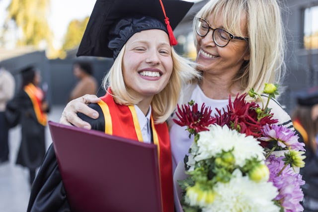 A joyful graduate in a cap and gown hugging an older woman while holding flowers at a graduation cer...