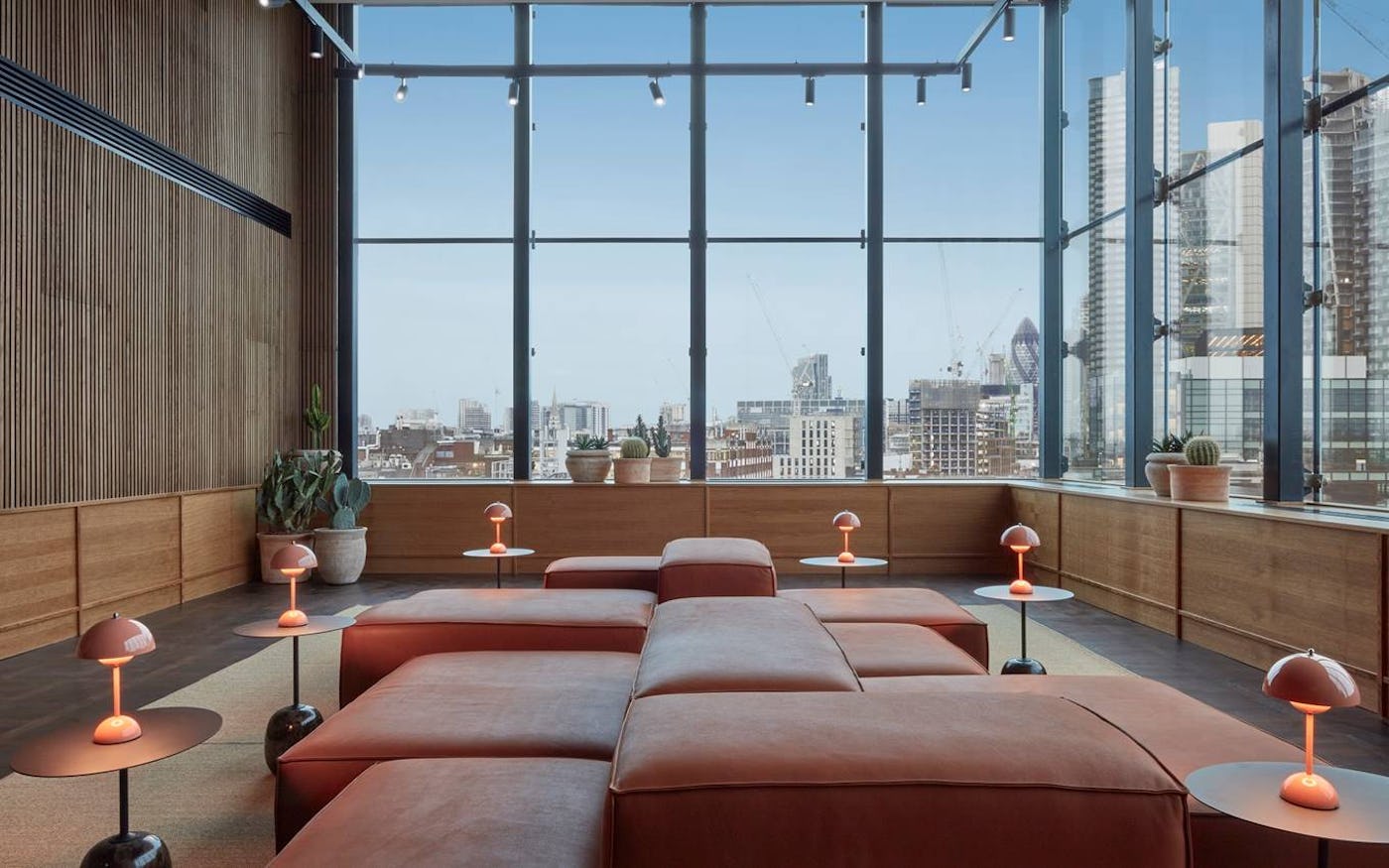Modern lounge with large brown sectional sofa and unique lamps, set against floor-to-ceiling windows overlooking a cityscape.