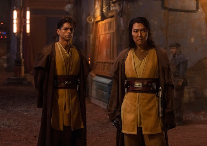 Two Jedi characters in traditional robes stand focused in a dim, dusty street, exuding a sense of readiness and concern.