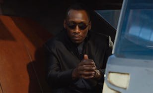 A man in sunglasses and a black jacket sits inside a car, clasping his hands together in a thoughtful pose.