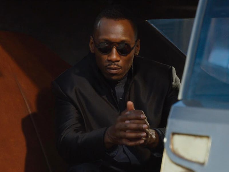 A man in sunglasses and a black jacket sits inside a car, clasping his hands together in a thoughtfu...