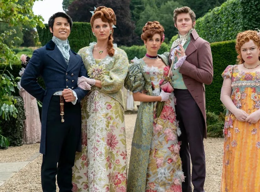 Five people in historical costumes stand in a garden; two men and three women wearing elaborate, flo...