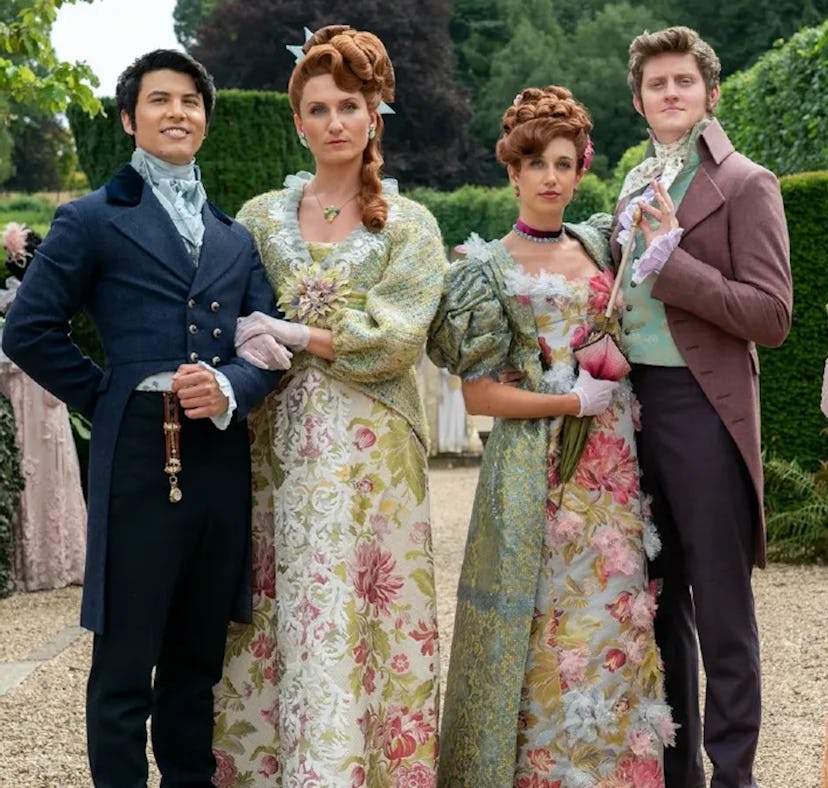 Five people in historical costumes stand in a garden; two men and three women wearing elaborate, flo...